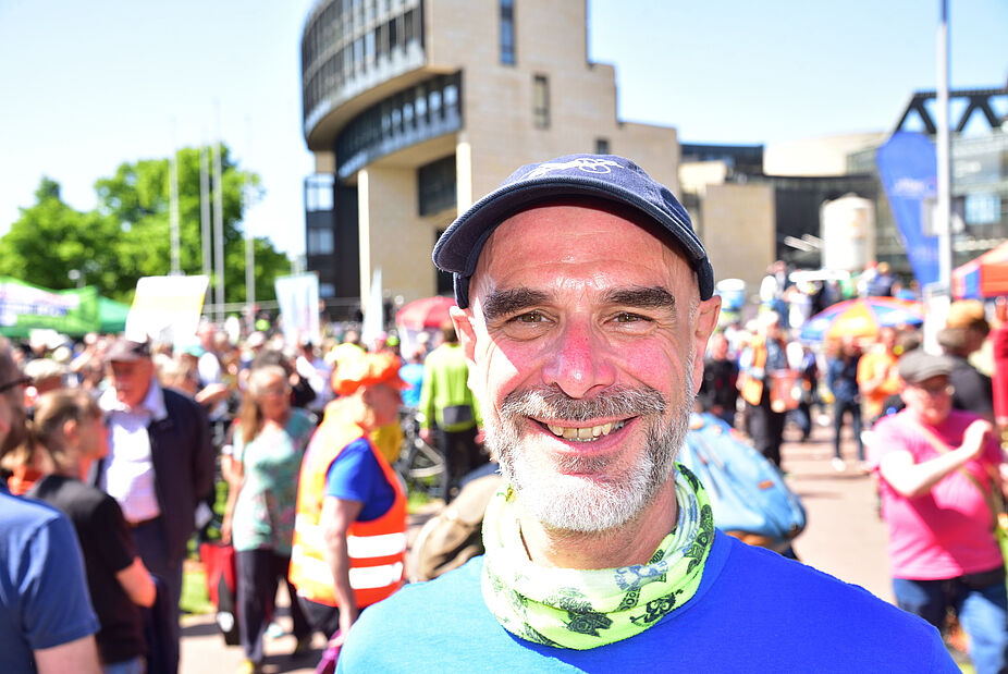 Axel Fell, Landesvorsitzender des ADFC NRW  Portrait Axel Fell, Landesvorsitzender des ADFC NRW, mit blauem T-Shirt, buntem Halstuch und ADFC-Kappe vor Menschenmenge und Landtag NRW im Hintergrund bei sonnigem Wetter. .