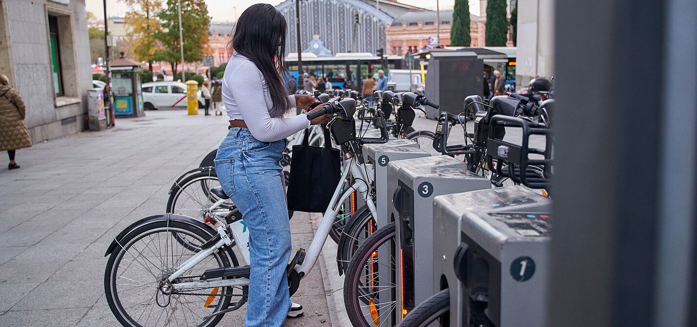 Frau entleiht Fahrrad an Bike-Sharing-Station Eine junge Frau mit langen dunklen Haaren, weißem Oberteil, Jeans und schwarzer Umhängetasche steht an einer Bike-Sharing-Station und entnimmt ein weißes Leihfahrrad. Die Station verfügt über mehrere nummerierte Stellplätze mit grauen Dockingelementen. Im Hintergrund sind ein belebter Stadtplatz, historische Gebäude, Passanten und ein Bahnhofsgebäude zu sehen.